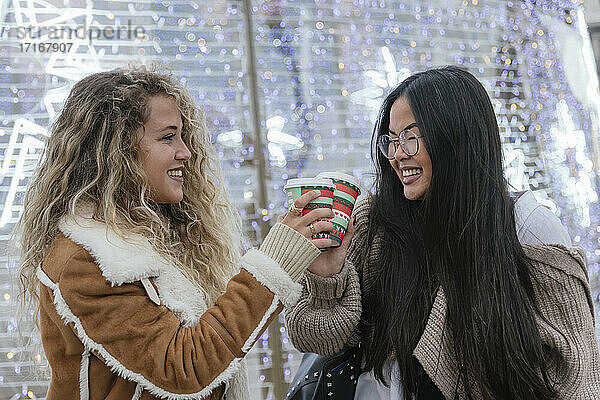 Beautiful female friends toasting coffee cups against illuminated Christmas lights