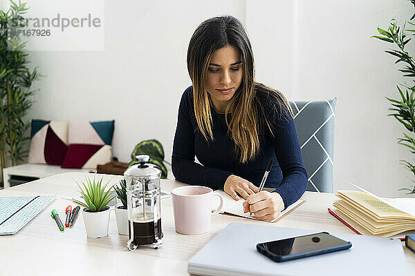 Junge Studentin schreibt in einem Buch  während sie im Wohnzimmer an der Wand sitzt