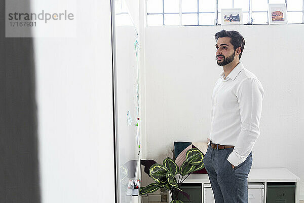 Lächelnder männlicher Unternehmer mit Händen in den Taschen  der im Büro auf ein Whiteboard schaut
