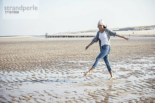 Unbeschwerte junge Frau mit Strickmütze  die auf nassem Sand am Strand spazieren geht