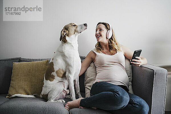 Schwangere Mutter hört Musik über Kopfhörer  während sie mit ihrem Hund zu Hause auf dem Sofa sitzt