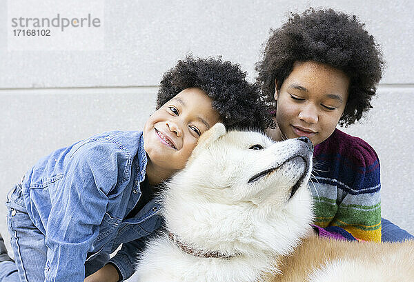Curly Haar Bruder und Schwester spielen mit Hund  während gegen graue Wand sitzen