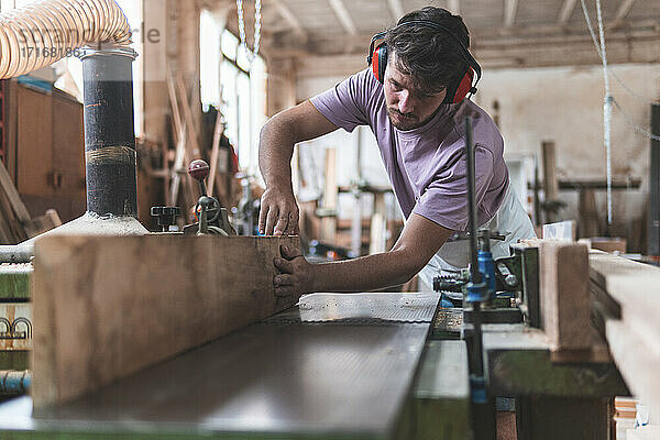 Young male craftsperson holding wooden plank on workbench while working in workshop