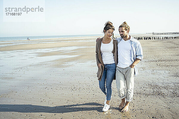 Junges Paar mit Händen in den Taschen bei einem Strandspaziergang an einem sonnigen Tag