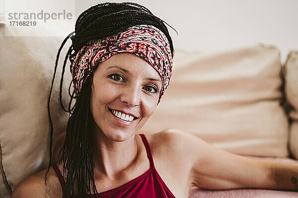 Lächelnde Frau mit Kopftuch starrt  während sie zu Hause auf dem Sofa sitzt