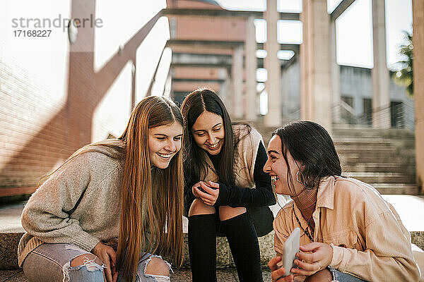 Fröhliches Teenager-Mädchen zeigt Freunden ihr Smartphone  während sie auf einer Treppe in der Stadt sitzt