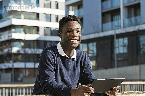 Smiling young man with digital tablet leaning on railing against building during sunny day