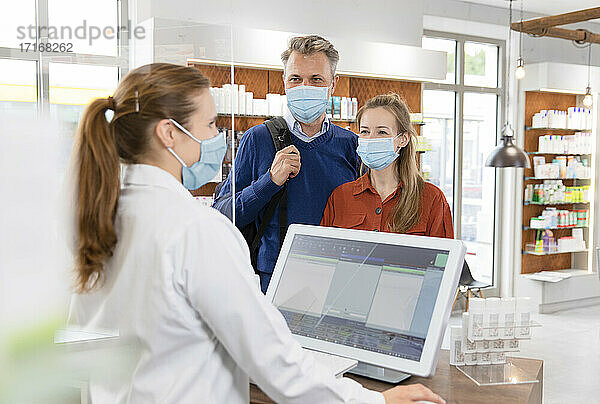 Female cashier with customers in protective face masks at pharmacy checkout