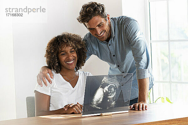 Lächelndes Paar prüft Ultraschallbild auf transparentem Bildschirm auf dem Tisch zu Hause