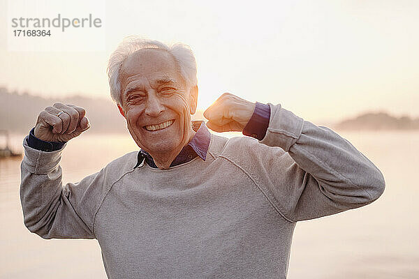 Lächelnder älterer Mann  der seinen Bizeps zeigt  während er gegen den Himmel bei Sonnenaufgang steht