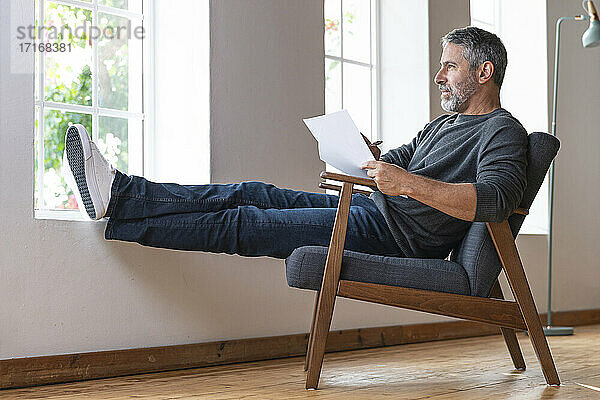 Reifer Geschäftsmann  der wegschaut  während er mit Papier auf einem Sessel zu Hause sitzt