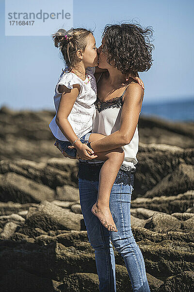 Affectionate mother kissing daughter while standing on flysch against blue sky