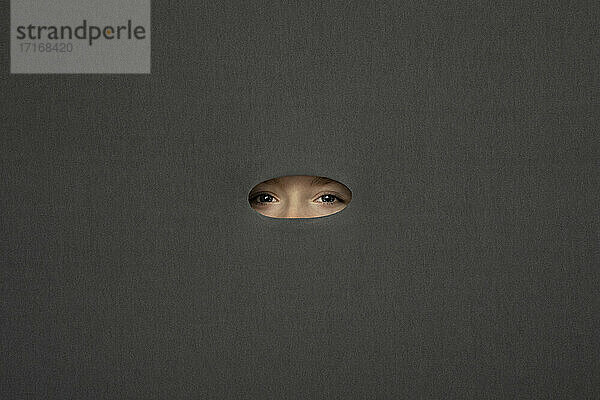 Eyes of teenage girl peeking through hole in gray background