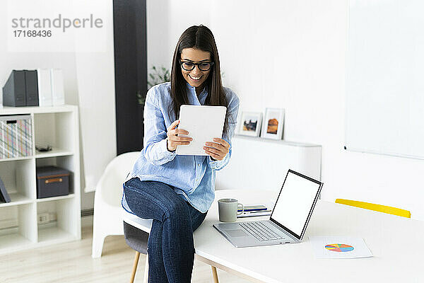 Lächelnde Geschäftsfrau  die an einer Telefonkonferenz auf einem digitalen Tablet teilnimmt  während sie im Büro am Laptop sitzt