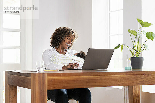 Lächelnde Frau mit Baby im Arm bei der Arbeit am Laptop im Büro zu Hause