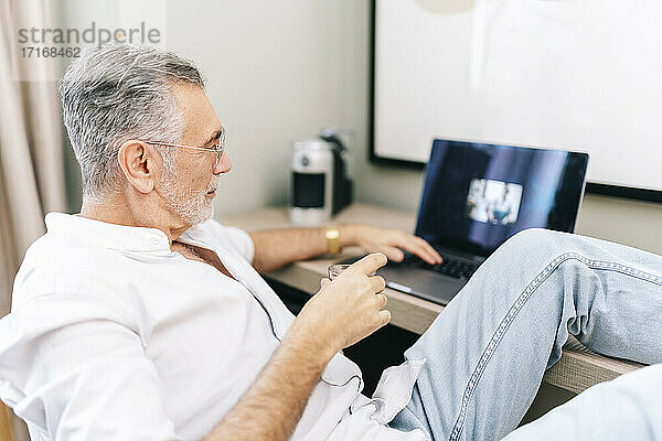 Mature man using laptop with feet up at desk while having coffee in hotel room