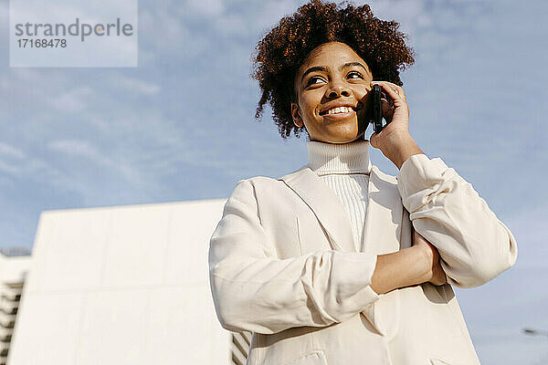 Junge Frau in weißem Mantel  die über ein Mobiltelefon spricht  während sie vor dem Himmel steht