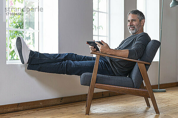 Lächelnder Geschäftsmann  der ein digitales Tablet benutzt  während er zu Hause auf einem Sessel sitzt
