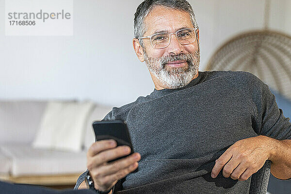 Lächelnder Geschäftsmann mit Brille  der ein Mobiltelefon benutzt  während er zu Hause sitzt
