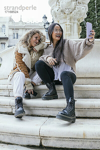 Cheerful female friends taking selfie through smart phone while sitting on steps by fountain