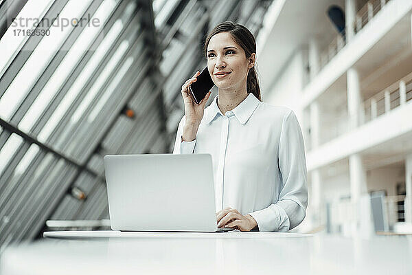 Lächelnde Unternehmerin  die mit einem Smartphone spricht  während sie im Flur an einem Laptop arbeitet