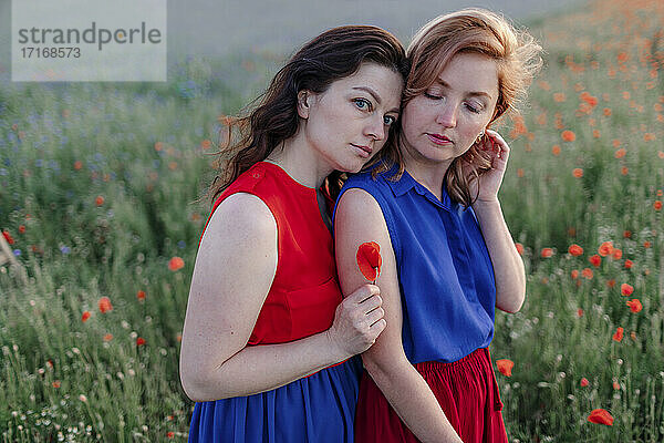 Woman holding flower while leaning on female friend's shoulder in poppy field