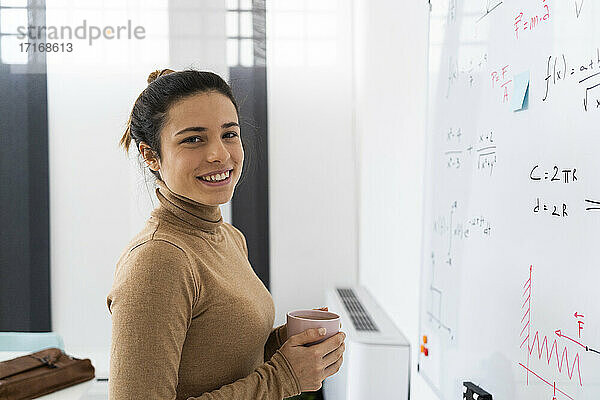 Lächelnde Frau hält eine Kaffeetasse  während sie zu Hause vor einem Whiteboard steht
