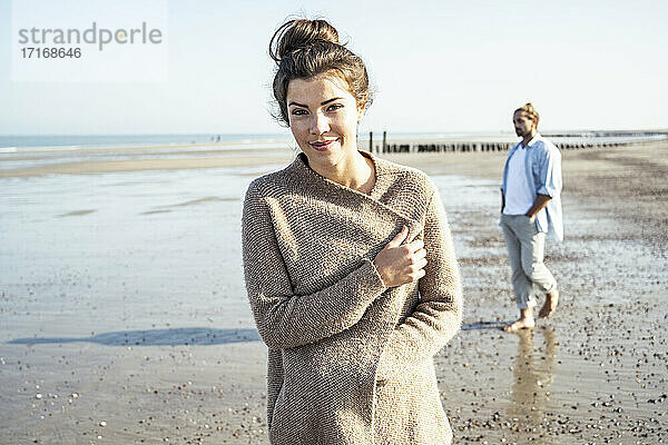 Junge Frau steht am Strand  während ihr Freund im Hintergrund am Wochenende spazieren geht