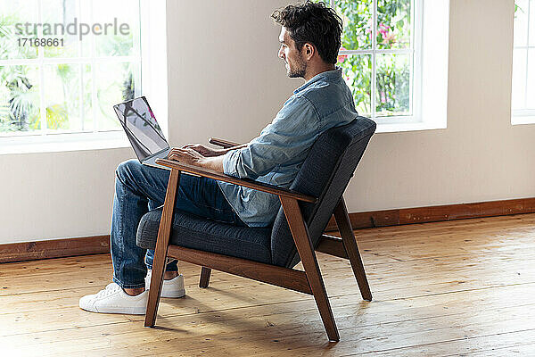 Mittlerer erwachsener Mann arbeitet am Laptop  während er zu Hause auf einem Sessel sitzt