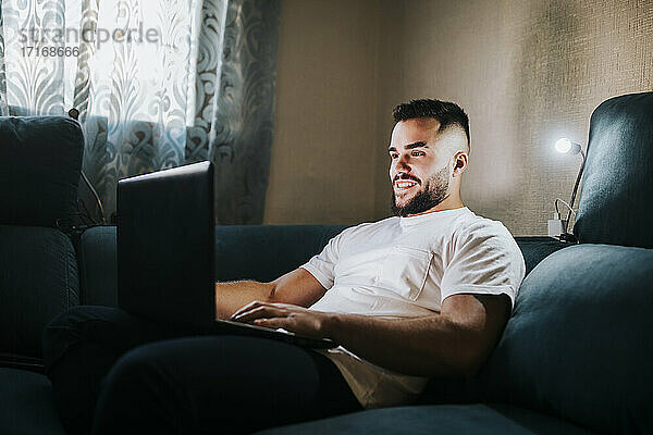 Lächelnder männlicher Unternehmer  der an einem Laptop arbeitet  während er auf der Couch im Wohnzimmer sitzt