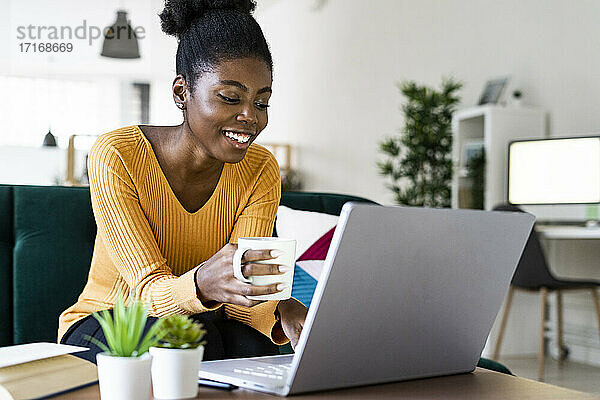 Happy Afro woman holding cup while using laptop at home