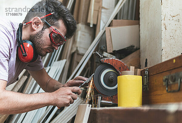 Concentrated male carpenter sharpening work tool on grinder in workshop