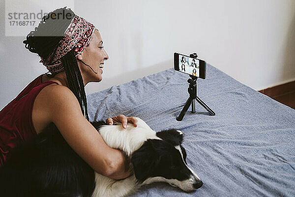 Lächelnde Frau im Gespräch mit Freunden per Videokonferenz über das Mobiltelefon  während sie mit ihrem Hund zu Hause auf dem Bett liegt