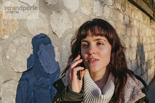 Nachdenkliche junge Frau  die bei Sonnenuntergang an einer Steinmauer mit ihrem Smartphone telefoniert