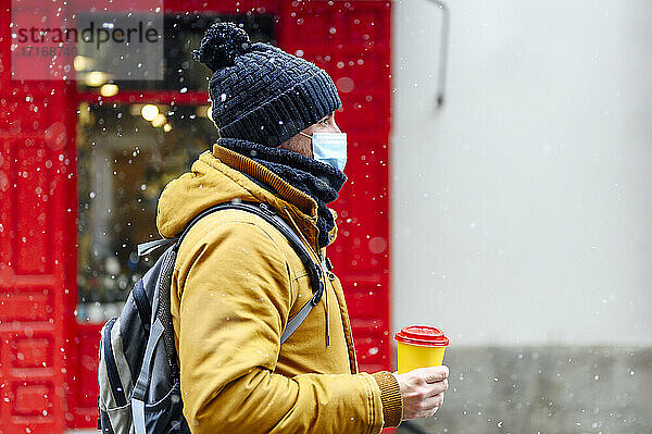 Man in warm clothing holding coffee cup while snowing during COVID-19