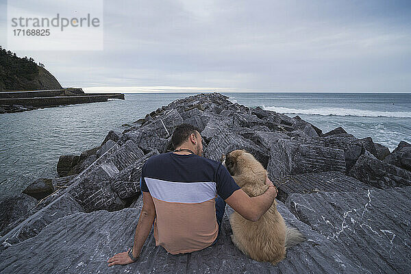 Man with arm around dog while sitting on rock against sky