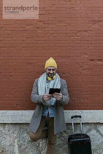 Mann in warmer Kleidung  der ein digitales Tablet benutzt  während er sich an eine Ziegelmauer lehnt