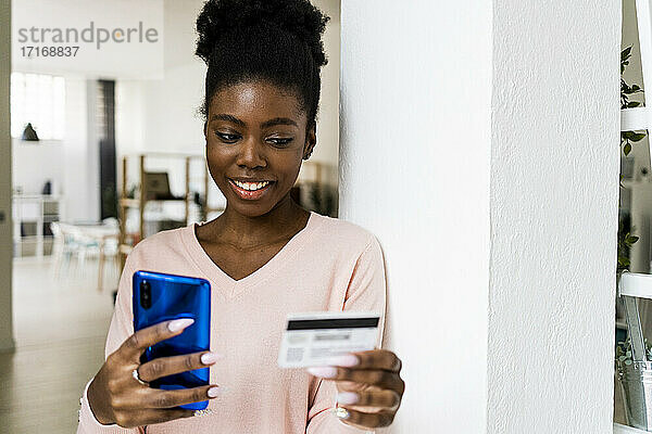 Frau mit Kreditkarte  die lächelt  während sie zu Hause ein Mobiltelefon benutzt