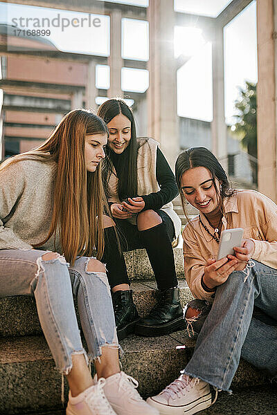 Lächelndes Teenager-Mädchen zeigt Freunden das Handy  während sie auf einer Treppe sitzt