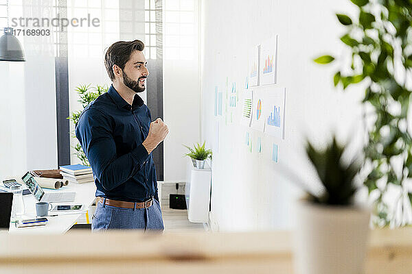 Cheerful male entrepreneur clenching fist while looking at strategies on wall in office