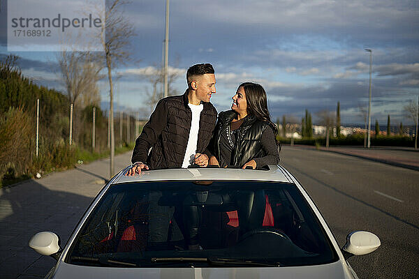 Junges Paar sieht sich an  während es auf dem Sonnendach eines Autos steht