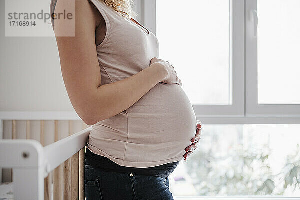 Schwangere Frau mit Händen auf dem Bauch am Fenster stehend zu Hause