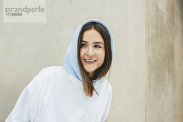 Smiling woman wearing hooded shirt looking away while standing against wall