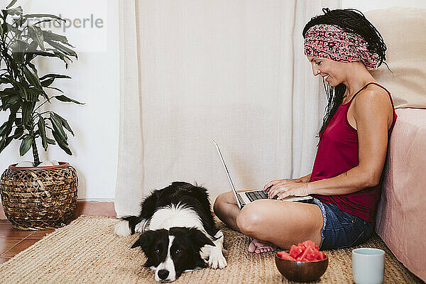 Frau arbeitet am Laptop  während sie bei Hund und Obstschale auf dem Teppich zu Hause sitzt