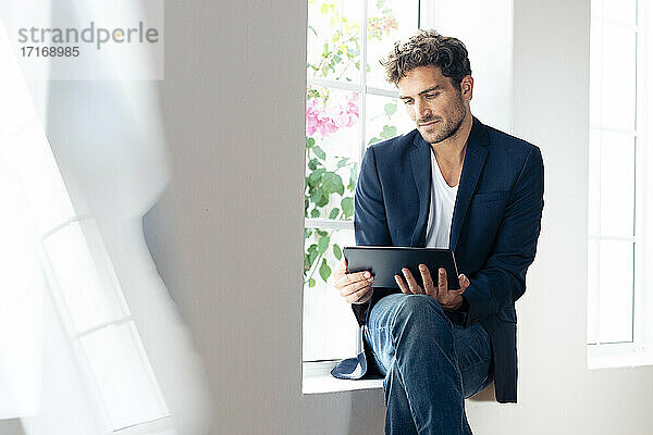 Geschäftsmann  der ein digitales Tablet benutzt  während er zu Hause auf der Fensterbank sitzt