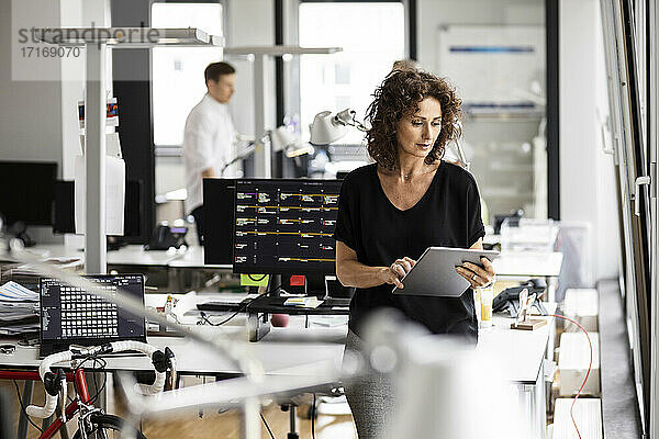 Geschäftsfrau  die ein digitales Tablet benutzt  während ein Kollege im Hintergrund in einem Großraumbüro steht
