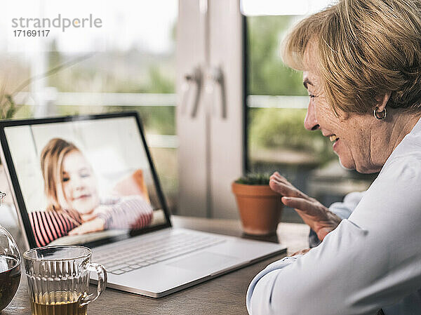 Lächelnde Großmutter  die ihrem Enkelkind bei einem Videogespräch über einen Laptop im Wohnzimmer zuwinkt