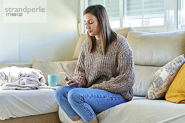 Female in warm clothing using smart phone on sofa at home