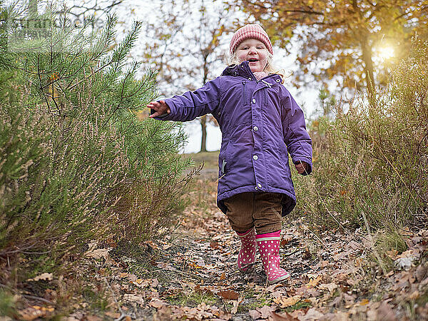 Lächelndes süßes Mädchen in lila Jacke  das inmitten von Pflanzen spazieren geht