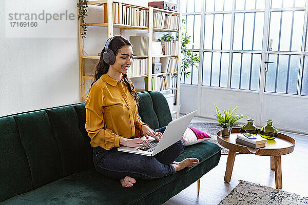 Woman wearing headphones using laptop while sitting on sofa at home
